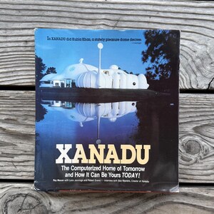 Puede incluir: Un libro titulado XANADU The Computerized Home of Tomorrow and How It Can Be Yours TODAY! por Roy Mason con Lane Jennings y Robert Evans. La portada muestra una casa blanca en forma de cúpula reflejada en agua oscura bajo un cielo azul.