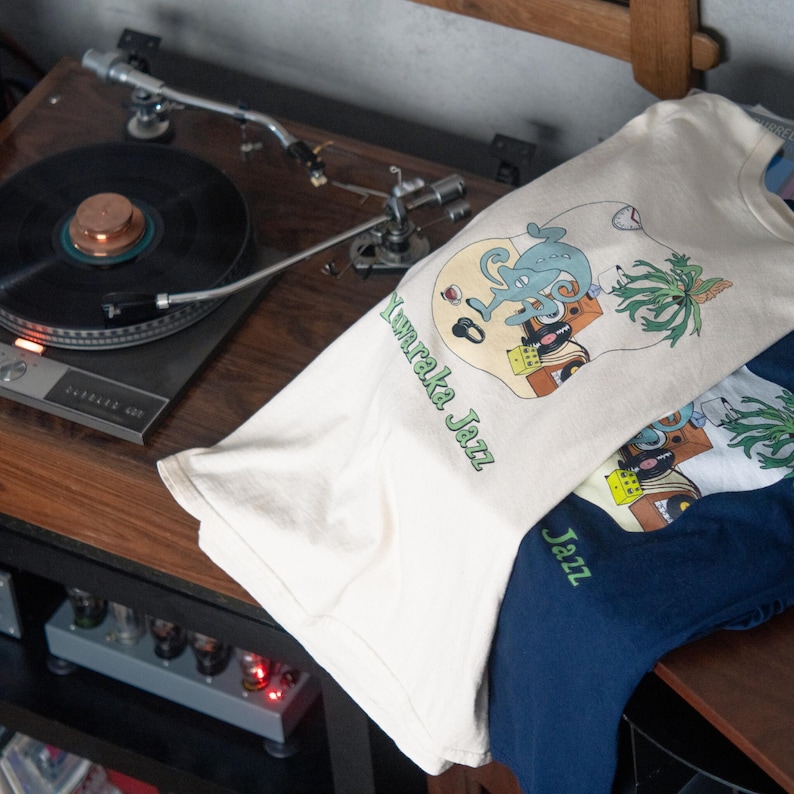 May include: A cream-colored t-shirt and a navy blue t-shirt, both featuring a graphic illustration of a blue chameleon, a palm tree, and musical equipment with the text "Yuraka Jazz" printed in green lettering on the cream shirt.