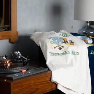 May include: An off-white t-shirt featuring a graphic illustration of a blue character, musical equipment, and the text Yawaraka Jazz in green. The design includes a small cup, headphones, and a plant, all set within a rounded shape.