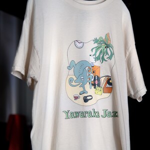 May include: A cream-colored short-sleeve t-shirt featuring a graphic print of a blue abstract creature, a clock, a plant, a speaker, a record player, headphones, and a cup. The text Yawaraka Jazz is printed in green at the bottom of the graphic.