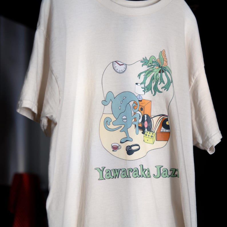 May include: A cream-colored short-sleeve t-shirt featuring a graphic print of a blue abstract creature, a clock, a plant, a speaker, a record player, headphones, and a cup. The text Yawaraka Jazz is printed in green at the bottom of the graphic.