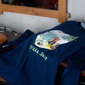 May include: A navy blue long-sleeve shirt featuring a graphic print of a light blue creature, a yellow die, a vinyl record, and a plant. The text "Yuraka Jazz" is printed in light green below the circular graphic on the front of the shirt.