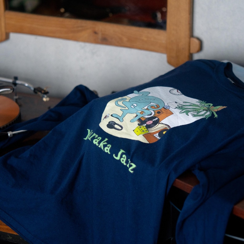 May include: A navy blue long-sleeve shirt featuring a graphic print of a light blue creature, a yellow die, a vinyl record, and a plant. The text "Yuraka Jazz" is printed in light green below the circular graphic on the front of the shirt.