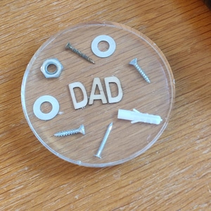 May include: A clear, round coaster with the word "DAD" spelled out with screws and washers. A ball of colorful rubber bands and a black remote control are also visible on a wooden surface.