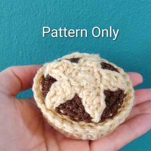 May include: A small, round, crocheted pie with a light beige crust and a dark brown filling. The crust features a star-shaped lattice design on top. The text 'Pattern Only' is printed in white above the crocheted item.