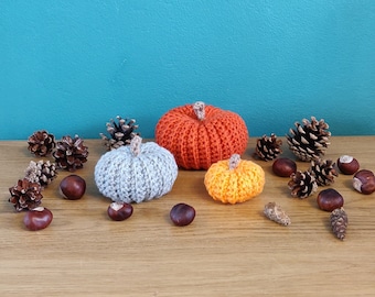 May include: A still-life arrangement featuring three crocheted pumpkins in orange, gray, and yellow, with pine cones and chestnuts. The pumpkins are on a wooden surface against a teal background. The pumpkins vary in size.
