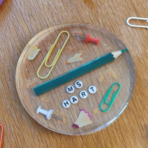 May include: A clear resin coaster with the name "MS HART" spelled out in letter beads, surrounded by a green pencil, paperclips, push pins, and a binder clip. A purple pencil and other office supplies are scattered around the coaster on a wooden surface.