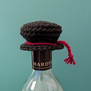 May include: A black crocheted bottle stopper with a red tassel. The bottle stopper is on top of a clear glass wine bottle with a black label that says "HARDYS".