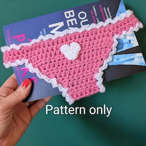 May include: A pink crocheted bookmark shaped like underwear with a white heart applique in the center and a white ruffled border. The item is placed over a book with a blue cover that has the text "OUR BE M" visible on the front.