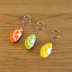 May include: Three crocheted fruit keychains with silver key rings. The keychains are shaped like a lime, an orange, and a lemon. The fruit are crocheted in shades of green, orange, and yellow.