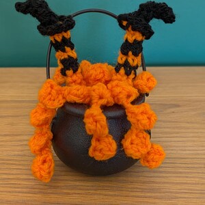 Pu&ograve; includere: Una decorazione a tema Halloween con un piccolo calderone nero con gambe da strega all'uncinetto arancioni e nere e un manico nero. Le gambe sono arancioni con strisce nere e cappelli da strega neri. Il calderone &egrave; su una superficie di legno.