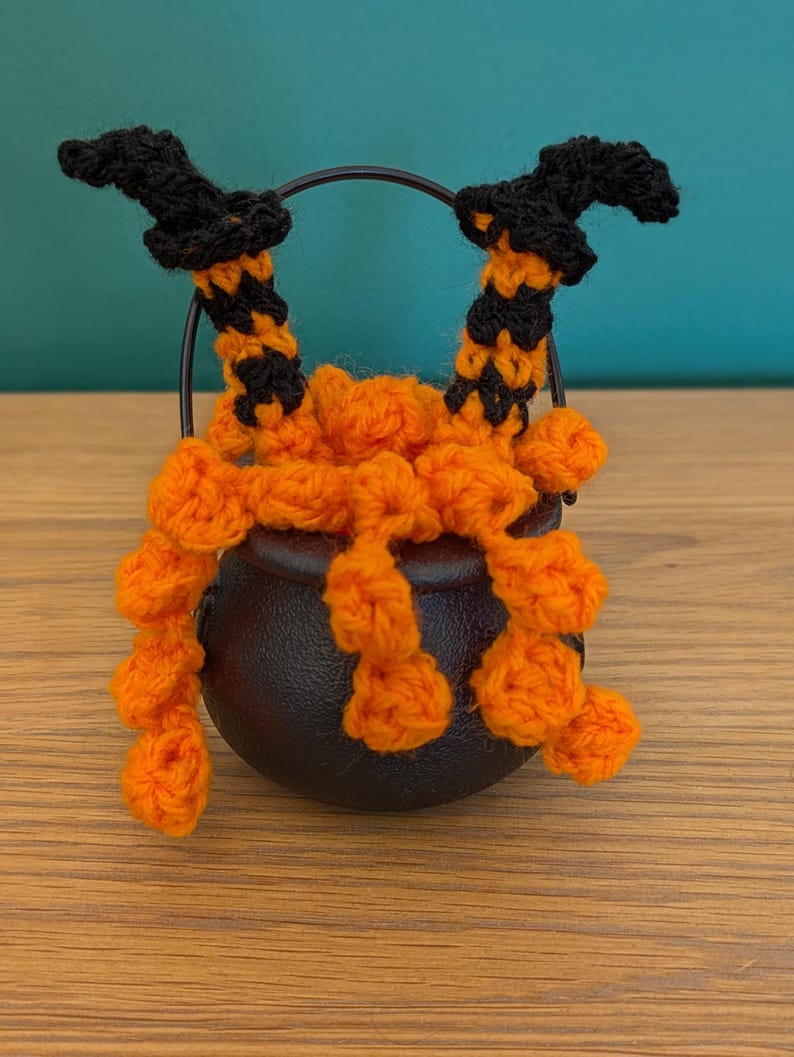 Pu&ograve; includere: Una decorazione a tema Halloween con un piccolo calderone nero con gambe da strega all'uncinetto arancioni e nere e un manico nero. Le gambe sono arancioni con strisce nere e cappelli da strega neri. Il calderone &egrave; su una superficie di legno.
