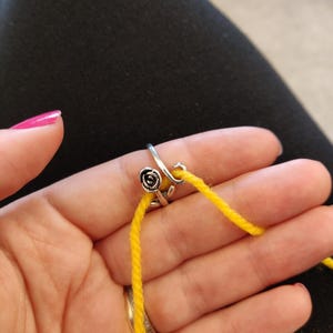Könnte beinhalten: Ein silberner Garnführungsring mit Rosenmuster hält gelbes Garn. Der Ring wird am Finger getragen, wobei sich das Garn über die Hand erstreckt. Der Hintergrund ist ein dunkler Stoff.