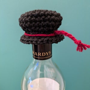 K&ouml;nnte beinhalten: Ein schwarzer, geh&auml;kelter Flaschenverschluss mit einem roten Band und einem langen roten Quasten. Der Flaschenverschluss befindet sich auf einer durchsichtigen Glasweinflasche mit dem Etikett "HARDYS".