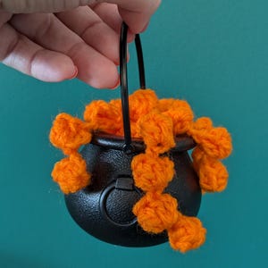 Pu&ograve; includere: Un piccolo calderone nero con un manico nero &egrave; riempito di pompon di filato arancione brillante. Il calderone &egrave; rotondo con una superficie strutturata. Lo sfondo &egrave; di colore turchese.