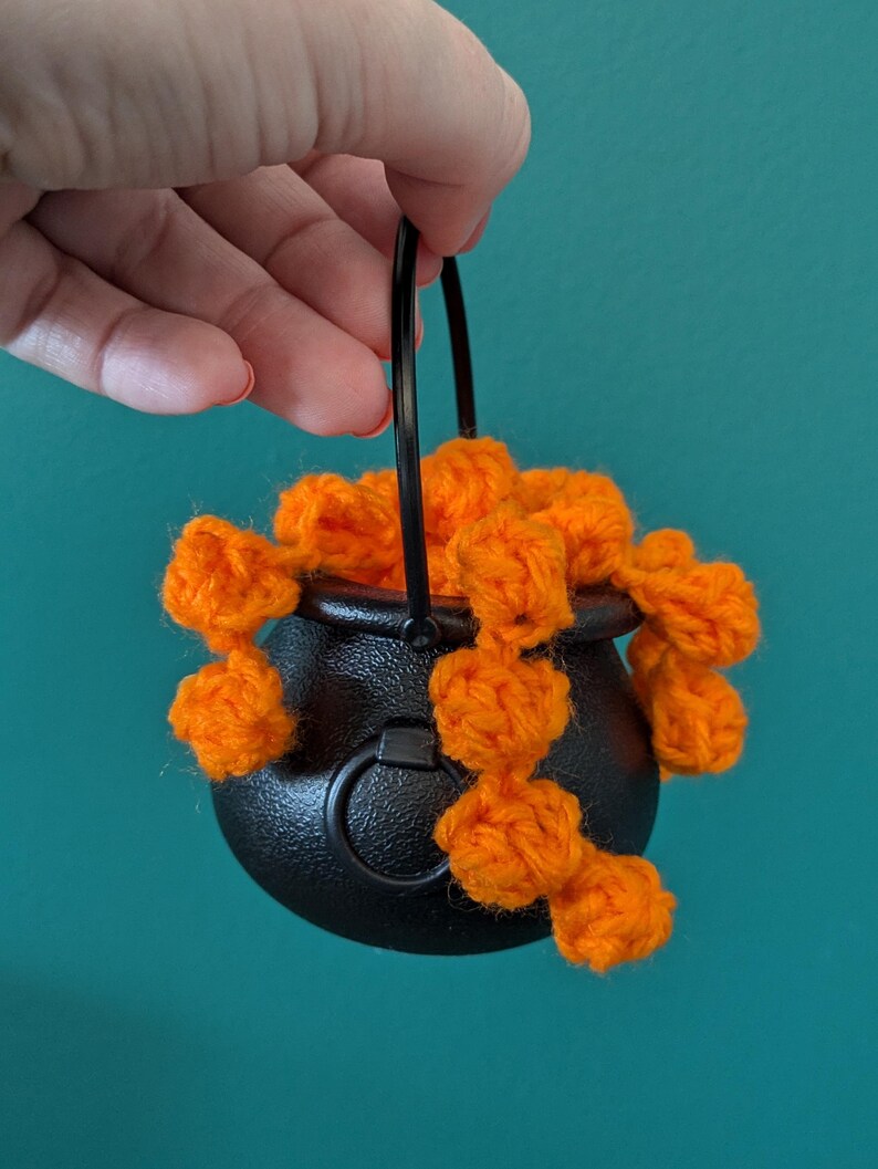 Pu&ograve; includere: Un piccolo calderone nero con un manico nero &egrave; riempito di pompon di filato arancione brillante. Il calderone &egrave; rotondo con una superficie strutturata. Lo sfondo &egrave; di colore turchese.