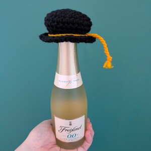 May include: A small bottle of Freixenet 0.0 alcohol-free sparkling wine topped with a black crocheted graduation cap. The cap features a yellow yarn tassel hanging from the side. The bottle has a white label with black text and gold accents.