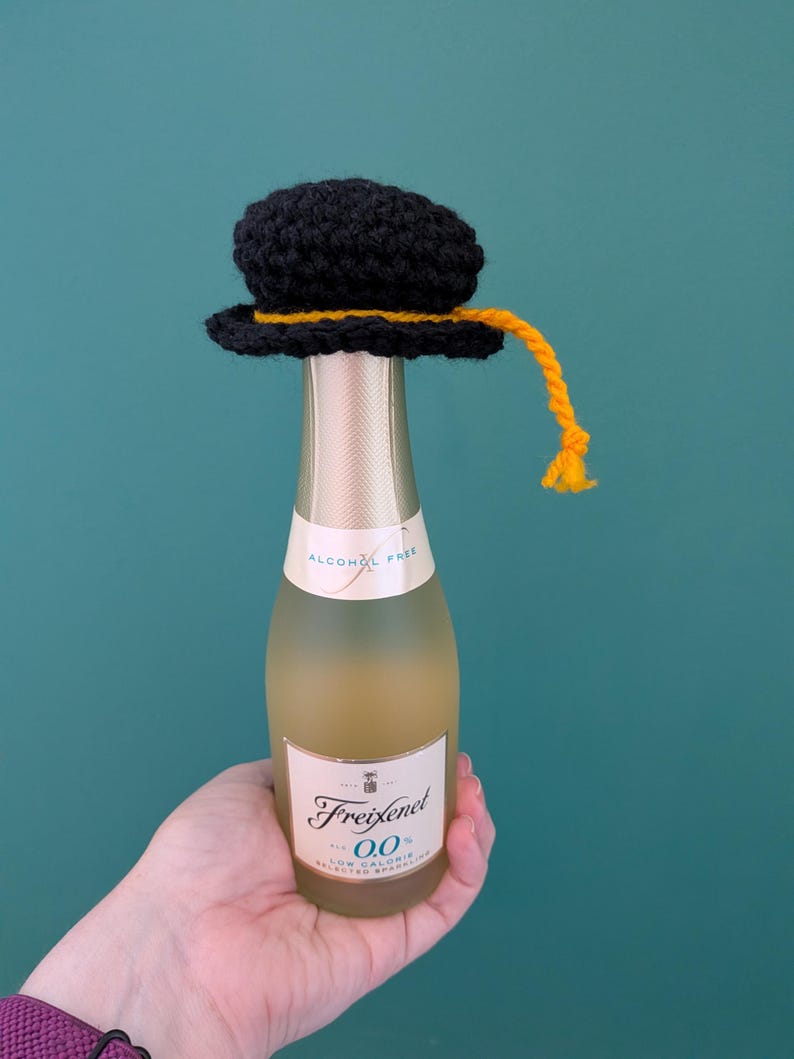 May include: A small bottle of Freixenet 0.0 alcohol-free sparkling wine topped with a black crocheted graduation cap. The cap features a yellow yarn tassel hanging from the side. The bottle has a white label with black text and gold accents.