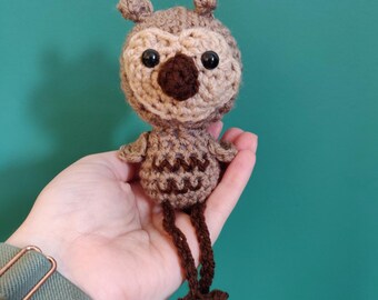 May include: A handmade crochet owl doll in shades of brown and tan. The owl has large black eyes, a brown beak, and small ear tufts. The owl's body features a zig-zag pattern and long, dangling legs with claw-like feet. The doll is held in a hand against a teal background.