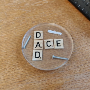 May include: A clear, round resin coaster with the word "DAD ACE" spelled out using Scrabble tiles. Two screws and a wall anchor are embedded in the resin. A black remote control is in the background.