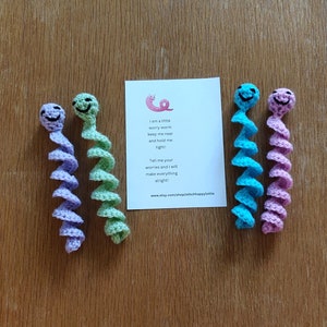 May include: Four crocheted worry worms in different colours: purple, green, blue, and pink. Each worm has a smiling face and is curled into a spiral shape. The worms are arranged in a row on a wooden surface. A card with a poem about worry worms is in the centre of the image. The poem reads: "I am a little worry worm keep me near and hold me tight! Tell me your worries and I will make everything alright!" The card also includes a website address: www.etsy.com/shop/stitchhappylottie