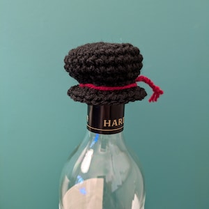 K&ouml;nnte beinhalten: Ein schwarzer, geh&auml;kelter Flaschenverschluss mit einem roten Band. Der Flaschenverschluss befindet sich auf einer durchsichtigen Glasflasche mit einem schwarzen Etikett, auf dem "HARDY'S" steht.
