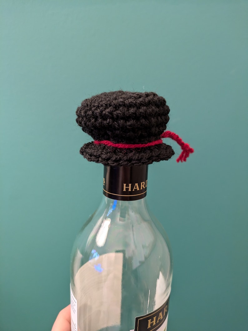 K&ouml;nnte beinhalten: Ein schwarzer, geh&auml;kelter Flaschenverschluss mit einem roten Band. Der Flaschenverschluss befindet sich auf einer durchsichtigen Glasflasche mit einem schwarzen Etikett, auf dem "HARDY'S" steht.