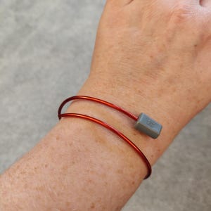 May include: A red metal bangle bracelet with a gray square charm. The charm has the letters "EG" on it.