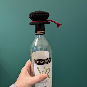 K&ouml;nnte beinhalten: Eine klare Glasflasche Hardy's VR Chardonnay Wein mit einem schwarzen geh&auml;kelten Flaschenverschluss mit einem roten Band.