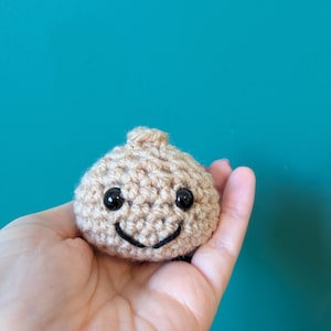Könnte beinhalten: Ein kleines, rundes, beiges Häkel-Plüschtier in Form einer Knoblauchknolle. Es hat zwei glänzende schwarze Kunststoffperlen als Augen und ein einfaches, mit schwarzem Garn aufgesticktes Lächeln. Die Struktur ist durch die Häkelarbeit knotig.