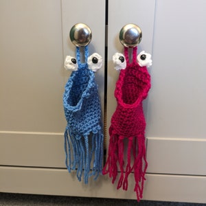 Könnte beinhalten: Zwei handgefertigte, gehäkelte Türgriffe in Monsterform. Einer ist blau, der andere rosa, beide mit weißen und schwarzen Augendetails und Fransen. Sie hängen an silbernen Knöpfen an einem grauen Schrank.
