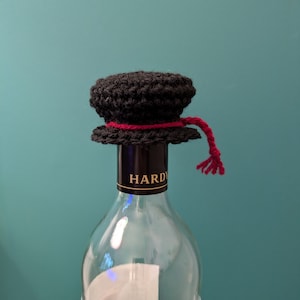 K&ouml;nnte beinhalten: Ein schwarzer, geh&auml;kelter Flaschenverschluss mit einem roten Band und einer Quaste. Der Flaschenverschluss befindet sich auf einer durchsichtigen Glasweinflasche mit einem schwarzen Etikett, auf dem "HARDY" steht.