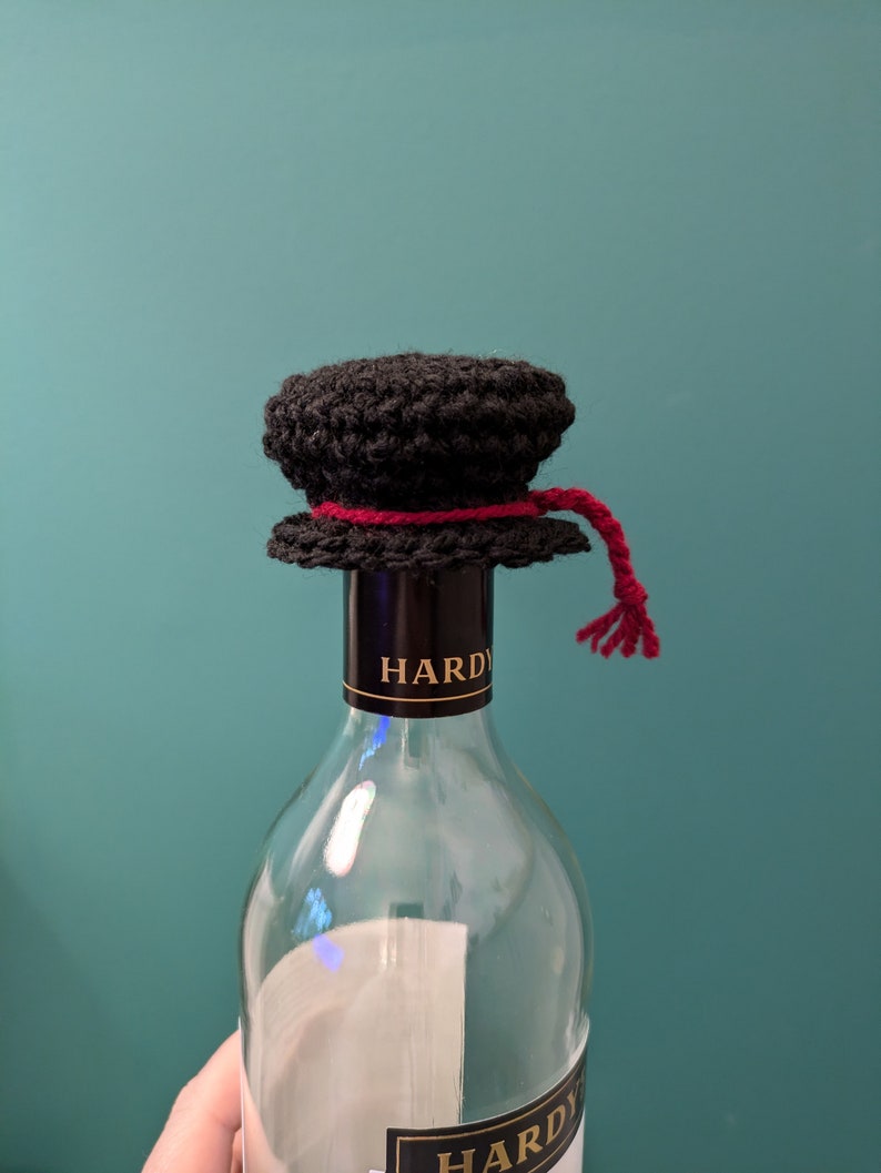 K&ouml;nnte beinhalten: Ein schwarzer, geh&auml;kelter Flaschenverschluss mit einem roten Band und einer Quaste. Der Flaschenverschluss befindet sich auf einer durchsichtigen Glasweinflasche mit einem schwarzen Etikett, auf dem "HARDY" steht.
