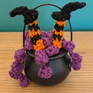Pu&ograve; includere: Una stravagante decorazione di Halloween con un calderone nero da cui fuoriesce filo viola. Due gambe da strega all'uncinetto, con cappelli neri e calze a righe arancioni e nere, spuntano dal calderone. Il calderone &egrave; nero.