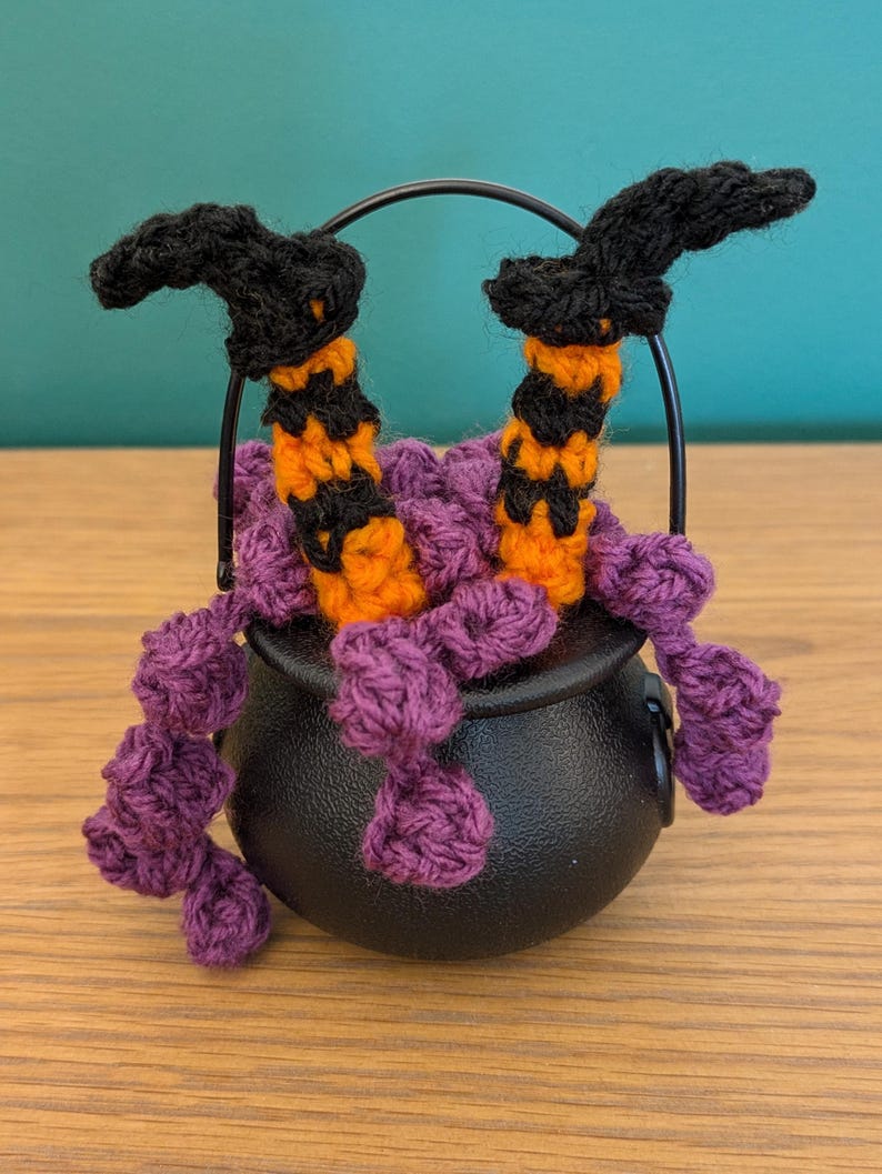 Pu&ograve; includere: Una stravagante decorazione di Halloween con un calderone nero da cui fuoriesce filo viola. Due gambe da strega all'uncinetto, con cappelli neri e calze a righe arancioni e nere, spuntano dal calderone. Il calderone &egrave; nero.