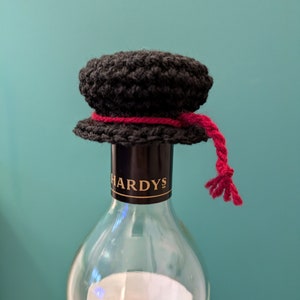 K&ouml;nnte beinhalten: Ein schwarzer, geh&auml;kelter Flaschenverschluss mit einem roten Band und einem Quasten. Der Flaschenverschluss befindet sich auf einer klaren Glasweinflasche mit einem schwarzen Etikett, auf dem "HARDYS" steht.