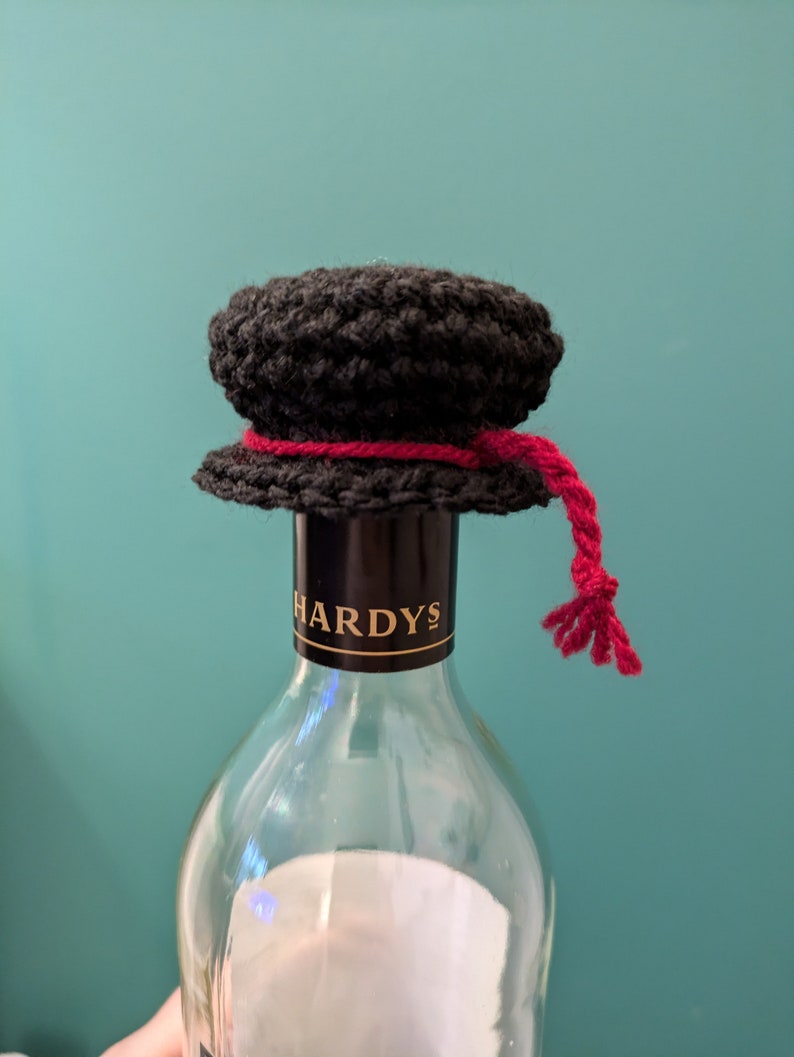 K&ouml;nnte beinhalten: Ein schwarzer, geh&auml;kelter Flaschenverschluss mit einem roten Band und einem Quasten. Der Flaschenverschluss befindet sich auf einer klaren Glasweinflasche mit einem schwarzen Etikett, auf dem "HARDYS" steht.