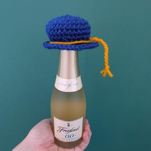 May include: A small, blue crocheted hat with a yellow yarn trim and a dangling yellow yarn tassel, placed on top of a Freixenet 0.0% alcohol-free sparkling wine bottle. The bottle features a cream-colored label with black text and gold accents.