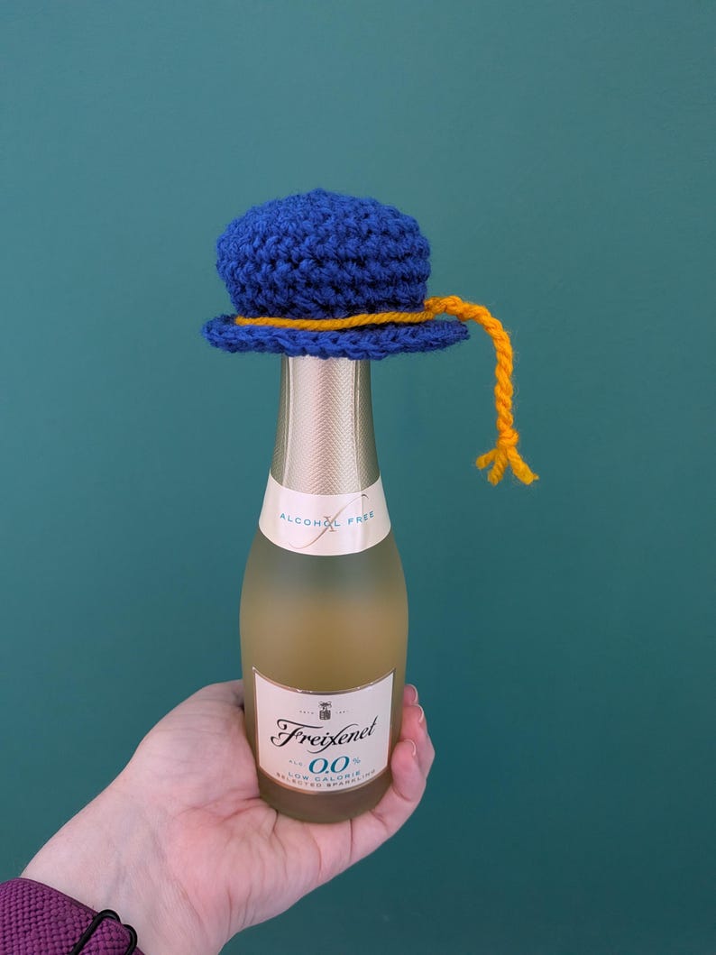 May include: A small, blue crocheted hat with a yellow yarn trim and a dangling yellow yarn tassel, placed on top of a Freixenet 0.0% alcohol-free sparkling wine bottle. The bottle features a cream-colored label with black text and gold accents.
