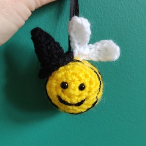 May include: A handmade crochet bee ornament with a smiling face, black eyes, and a black and white hat. The bee is yellow with black stripes and white wings. It hangs from a black ribbon.