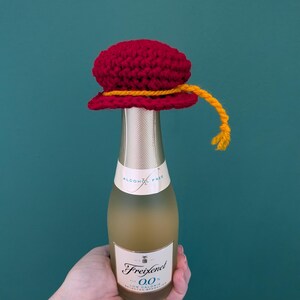 May include: A small glass bottle of Freixenet 0.0 alcohol-free sparkling wine topped with a handmade red crocheted hat. The hat features a decorative yellow yarn trim that hangs down the side. The bottle label displays the text Freixenet 0.0.