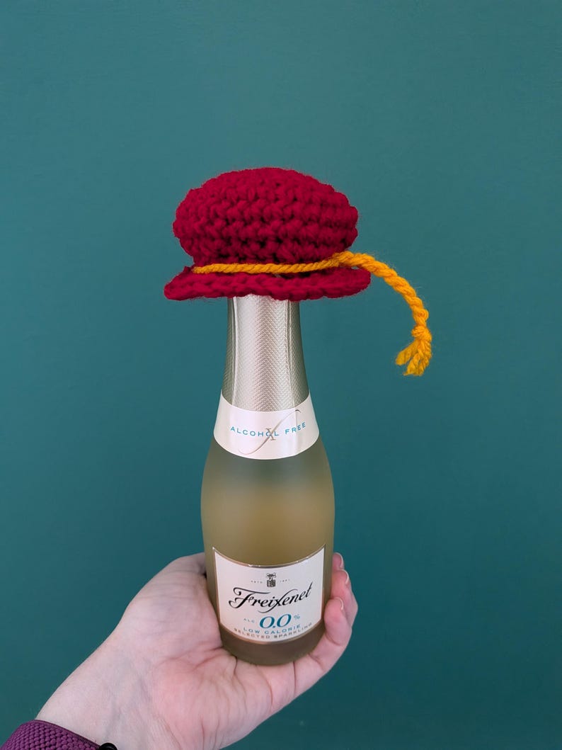 May include: A small glass bottle of Freixenet 0.0 alcohol-free sparkling wine topped with a handmade red crocheted hat. The hat features a decorative yellow yarn trim that hangs down the side. The bottle label displays the text Freixenet 0.0.