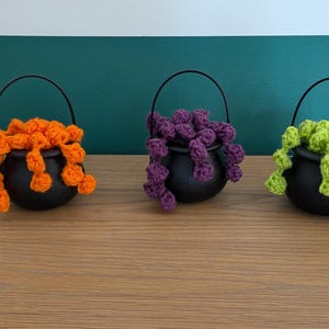 Pu&ograve; includere: Tre piccoli contenitori neri a forma di calderone con manici ad arco. Ogni calderone contiene un colore diverso di materiale all'uncinetto: arancione, viola e verde. I calderoni sono disposti su una superficie di legno contro uno sfondo turchese e bianco.