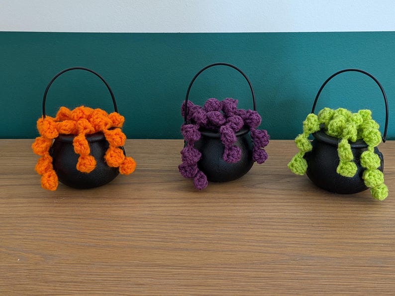 Pu&ograve; includere: Tre piccoli contenitori neri a forma di calderone con manici ad arco. Ogni calderone contiene un colore diverso di materiale all'uncinetto: arancione, viola e verde. I calderoni sono disposti su una superficie di legno contro uno sfondo turchese e bianco.