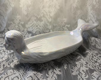 May include: A white ceramic dish shaped like a mermaid with an iridescent finish. The dish features a sculpted mermaid head at one end and a flared tail at the other, with a shallow, elongated basin in the center for holding small items.