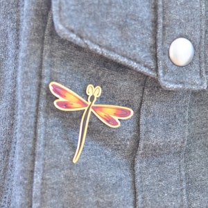 May include: A gold dragonfly pin with red and orange wings on a gray fabric background.