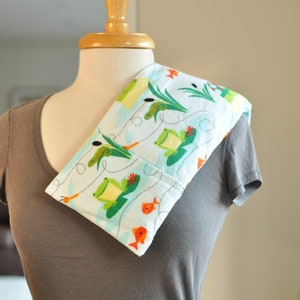 May include: A light blue fabric neck wrap with a white background and a pattern of green frogs, orange fish, and green lily pads. The wrap is on a grey shirt.