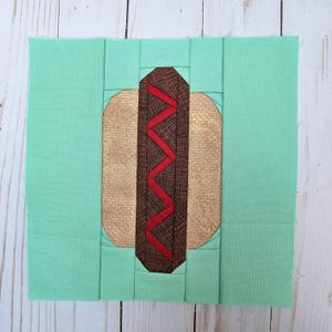 May include: A fabric block featuring a hot dog design. The hot dog has a brown bun, a brown and red zigzag-patterned sausage, and is set against a light green background. The block is square and appears to be handmade.