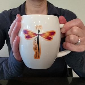 May include: White ceramic mug with a dragonfly design and the text "Skinny Bug Studio" in black and orange lettering.