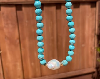 May include: A turquoise beaded necklace with a large, iridescent, oval-shaped pearl pendant. The necklace is displayed outdoors against a wooden fence and a bright blue sky. The beads are approximately 0.3 inches in diameter.
